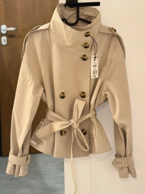 Beige trenchcoat med bälte och axelklaffar bershka - Helt ny oanvänd kort trench coat från Bershka. Säljer endast pga fel storlek. Lappen är kvar