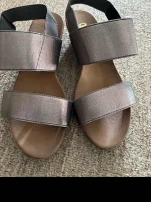Metallic kilklackade sandaler - Snygga sandaler med breda metallicgrå remmar och kilklack. Öppen tå och elastiskt band runt hälen för extra komfort. Perfekta för dig som vill ha både stil och höjd. Materialet ser ut att vara syntet med en glansig finish.
