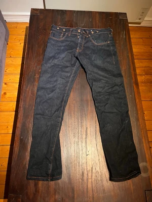 Ett par vintage jeans ifrån no.666 - Säljer ett par mörkblå jeans med raka ben och snygga kontrastsömmar. Jeansen har fem fickor, bälteshällor och coola broderade detaljer på bakfickorna. Klassisk knappgylf och slitstarkt denimtyg. Perfekta för en clean och stilren look. Kan även ta bort det svarta lädret innan jag fraktar. Nypris 2000kr, SIZE 33/34