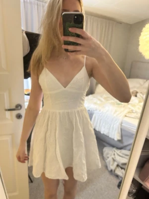 Vit kort spetsklänning med smala band - Söt vit kort klänning med smala axelband och v-ringning. Klänningen har en omlottliknande överdel och ett utställt kjolparti i spetsmaterial som ger en romantisk känsla. Perfekt för student, skolavslutning eller andra somriga tillställningar🩷