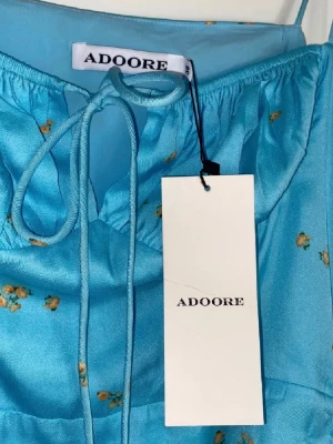 Blå blommig klänning från Adoore - Säljer min oanvända ljusblå klänning från Adoore med små blommor, smala axelband och snörning framtill. Klänningen har en figurnära passform och är tillverkad i ett glansigt material. Prislapp kvar!