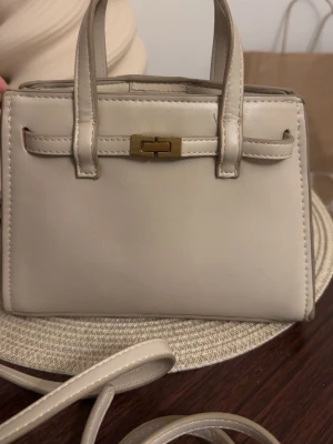 Beige väska - Snygg beige minihandväska i slätt konstläder med guldfärgade metalldetaljer och avtagbar axelrem. Väskan har två handtag och en dekorativ spänndetalj framtill. Perfekt för dig som gillar stilrena accessoarer med minimalistisk design. Aldrig använd. 