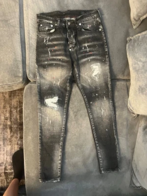 Svarta slimfit med slitningar från dsquared - Säljer ett par slim fit jeans med slitningar och färgstänk från Dsquared. Jeansen har en tvättad look, detaljerade slitningar på framsidan och färgstänk i vitt och rött. Klassisk femficksmodell och smal passform. Perfekta för en edgy streetstil.
