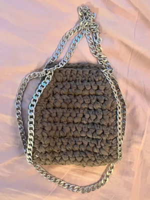Grå virkad handväska med kedja - Unik handväska i grått, virkad med tjockt garn och detaljerad med en bred silverfärgad kedja som axelrem. Väskan har en mjuk, fyrkantig form och ger en trendig och handgjord känsla. Perfekt accessoar för dig som gillar statement-väskor.