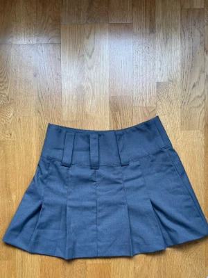 Grå minikjol med bred linning - Snygg grå minikjol med bred linning och markerade veck framtill. Kjolen har breda hällor och en stilren look som passar till många olika outfits. Perfekt för dig som gillar en enkel men trendig stil.