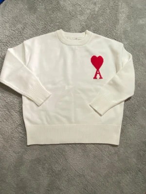 Vit stickad tröja med rött hjärta - Säljer en vit stickad tröja med rund hals och ribbade muddar. På bröstet finns ett rött hjärta och bokstaven A, vilket ger en unik och trendig look. Perfekt för dig som gillar stilrena plagg med en twist.