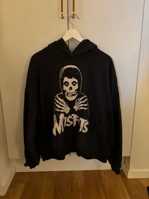 Svart Misfits hoodie med tryck - Säljer en svart hoodie från HM med stort vitt tryck på framsidan som föreställer ett skelett i hoodie och texten 'Misfits'. Klassisk huva och avslappnad passform. Perfekt för dig som gillar bandmerch och streetwear.