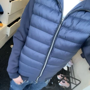 Mörkblå pufferjacka - Säljer en stilren mörkblå pufferjacka med dragkedja. Jackan har långa ärmar och en quiltad design som ger en modern look. Perfekt för kyligare dagar men även sommar kvällar . Är av märket ”tiffosi” och är köpt i Italien för 1500kr. Jackan är storlek s med en tightare passform!