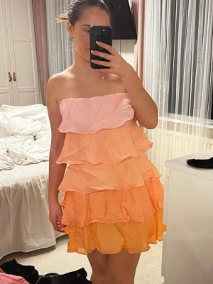 Pastellfärgad volangklänning axelbandslös - Säljer en helt ny söt med prislapp axelbandslös kortklänning i pastellfärger med flera lager volanger. Klänningen går i rosa, orange och gul och har en luftig, flowy känsla. Nypris: 1500