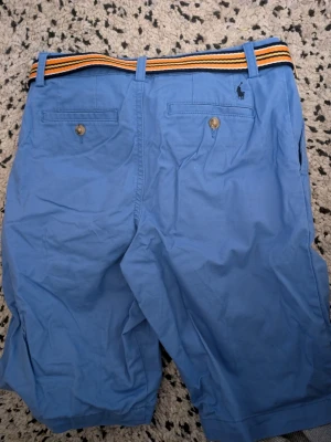 Blå shorts från Polo Ralph Lauren stl 18 - Snygga blå shorts från Polo Ralph Lauren med randigt bälte i orange, svart och vitt. Klassisk design med två bakfickor med knappar och broderad logga vid linningen. Perfekta för  mycketsommardagar och avslappnad stil. Nyskick står stl 18 men skulle säga passar 158-164