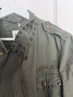 Grön parkajacka med nitar - Snygg grön parkajacka med coola nitar på krage och fickor. Jackan har axelklaffar, tryckknappar och dragkedja framtill. Står xxs men är oversized i modellen 
