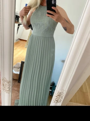 Ljusgrön långklänning med spets - Snygg ljusgrön långklänning med spetsdetaljer upptill och plisserad kjol. Klänningen har hög halsringning och är ärmlös, vilket ger en elegant och modern look. Perfekt för speciella tillfällen eller när du vill känna dig extra fin.