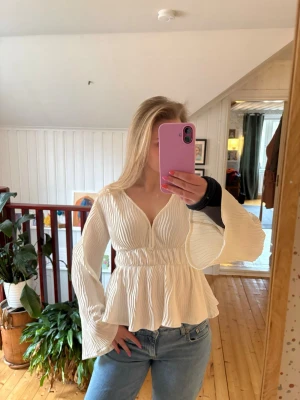 Vit plisserad blus med v-ringning - Trendig vit blus med plisserat tyg och markerad midja. Blusen har djup v-ringning och vida, långa ärmar. Perfekt att styla med jeans eller kjol för en snygg look. Materialet är lätt och luftigt, vilket gör den bekväm att bära.