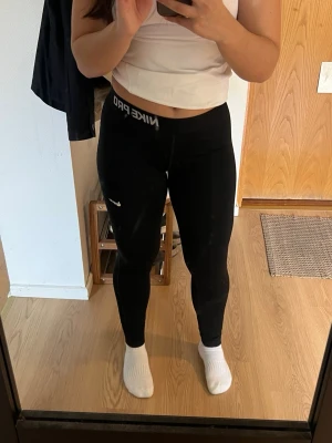 Svarta Nike Pro leggings - Svarta Nike Pro leggings med lite lägre midja och elastisk linning där det står 'NIKE PRO' i vitt. Swoosh-logga på benet är lite urtvättat tyvär och det finns även ett litet hål längre ner vid benet (se bild 3). Snygg och tight passform, perfekt för träning. 🥰
