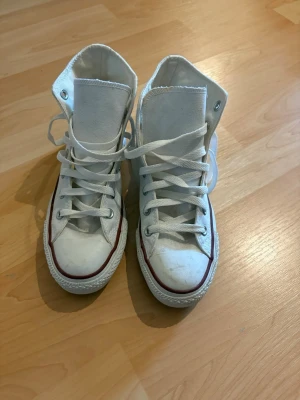 Vita Converse Chuck Taylor Highs - Klassiska vita Converse Chuck Taylor All Star high tops med vit canvas, vita snören och gummisula. Röd och svart rand längs sulan och ikonisk Converse-logga på sidan. Perfekta för en avslappnad och cool stil.