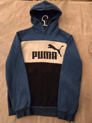 Blå och svart hoodie från Puma - Snygg hoodie från Puma i blått, vitt och svart med stor logga och text på bröstet. Klassisk huva och magficka framtill. Tillverkad i mjukt bomullsmaterial som är skönt att ha på sig. Perfekt för dig som gillar sportig och avslappnad stil.