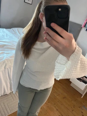 Vit långärmad topp med trumpetärm - Säljer en vit långärmad topp med strukturmönster och vida trumpetärmar. Toppen har en mjuk, stretchig känsla och sitter figurnära. Perfekt för dig som gillar unika detaljer och en stilren look. Super skön och perfekt nu i sommar samt vår. Fungerar bra hela året också. Säljer även de gröna jeansen i första bilden! 