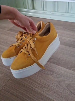 Gula platåsneakers i mocka - Säljer ett par snygga gula sneakers med platåsula och snörning. Skorna är gjorda i mocka och har en tjock vit sula som ger extra höjd. Perfekta för dig som vill sticka ut med färg och stil.