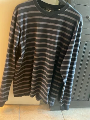 Randig långärmad tröja Mads Nørgaard - Snygg långärmad tröja från Mads Nørgaard med svarta och gråa horisontella ränder. Klassisk rund halsringning och mjukt material som passar perfekt till jeans eller chinos. Enkel men stilren design för en avslappnad look.
