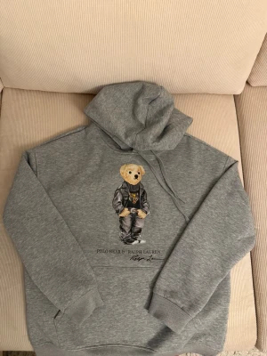 Grå hoodie Polo Bear Ralph Lauren - Snygg grå hoodie från Ralph Lauren med Polo Bear-tryck på bröstet. Klassisk huva med dragsko och magficka. Mjuk bomullsblandning och ribbade muddar. Perfekt för en chill och trendig look.