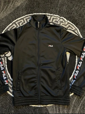 Svart FILA ziptröja med logga - Svarte ziptröja från FILA med hög krage och dragkedja. Tröjan har vita och röda FILA-loggor längs båda ärmarna samt en liten logga på bröstet. Tillverkad i ett glansigt, sportigt material och har ribbade muddar vid ärmslut och nederkant.