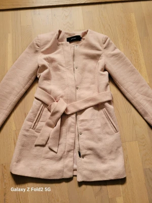 Ljusrosa kappa från Vero Moda - Stilren ljusrosa kappa från Vero Moda med knytbälte i midjan och dold dragkedja framtill. Kappan har rund halsringning, långa ärmar och två fickor. Perfekt för dig som gillar en enkel och elegant look.