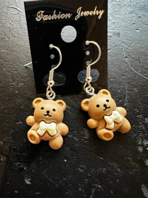 Söta björnörhängen med rosett - Unika örhängen med gulliga bruna björnar i plast, dekorerade med vita rosetter. Hängande modell med silvriga krokar. Perfekta för dig som vill ha en lekfull och trendig accessoar som sticker ut.