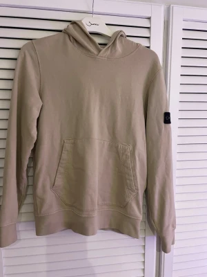Beige hoodie från Stone Island - Snygg beige hoodie från Stone Island med klassisk patch på vänster ärm. Hoodien har huva, känguruficka framtill och ribbade muddar. Tillverkad i mjuk bomull som känns skön mot huden. Perfekt för en avslappnad och trendig stil.