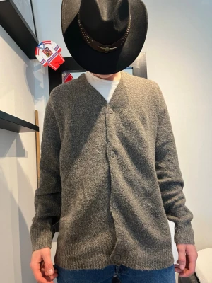 Gråmelerad kofta med knappar - Mysig gråmelerad kofta med v-ringning och knäppning framtill. Koftan har långa ärmar och ribbade muddar vid ärmslut och nederkant. Perfekt att slänga över en t-shirt för en avslappnad look. Modelen är 182 och väger 75 kg så är man lite större så är den perfekt 