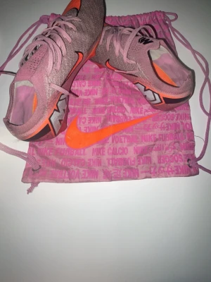 Nike rosa fotbollsskor med orange detaljer - Säljer ett par snygga rosa Nike fotbollsskor med orange swoosh och detaljer. Skorna har en strömlinjeformad design och är tillverkade i ett lätt syntetmaterial för bästa känsla på planen. Sulan har dobbar för bra grepp på gräs. Perfekta för dig som vill sticka ut!