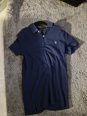 Marinblå pikétröja från Gant - Klassisk marinblå pikétröja från Gant med korta ärmar och krage. Tröjan har knappar framtill och en liten logga broderad på bröstet. Perfekt till jeans eller shorts för en clean och stilren look.