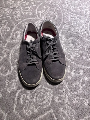 Grå mockasneakers  - Stilrena mörkgrå sneakers i mjuk mocka med vita sulor och klassisk låg profil. Skorna har matchande mörkgrå snören och en enkel, minimalistisk design som passar till det mesta. Insidan är ljus med en touch av rött vid hälen.