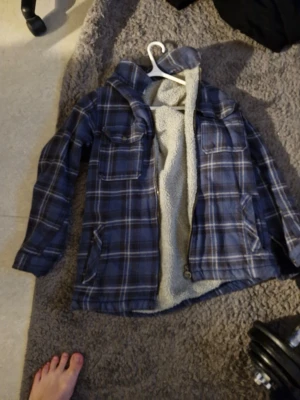 Blå rutig overshirt med teddyfoder - Säljer en blå och svart rutig overshirt med mysigt vitt teddyfoder på insidan. Jackan har två bröstfickor med lock, sidofickor och dragkedja framtill. Perfekt för lager på lager och chill stil.