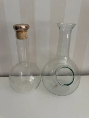 Två karaffer  i glas - Säljer två karaffer  i klart glas. Den ena har en rund botten och lång hals med metallock som har en symbol ingraverad. Den andra flaskan har också rund botten och bredare hals utan lock. Perfekta som dekoration eller som vattenkaraffer .