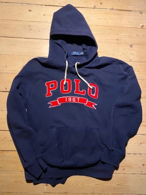 Marinblå hoodie från Polo Ralph Lauren - Snygg marinblå hoodie från Polo Ralph Lauren med stor röd POLO-logga och 1967 broderat framtill. Klassisk känguruficka, vita dragsnören och mjukt bomullsmaterial. Den passar  som en storlek L då den passar mig som generellt bär L.