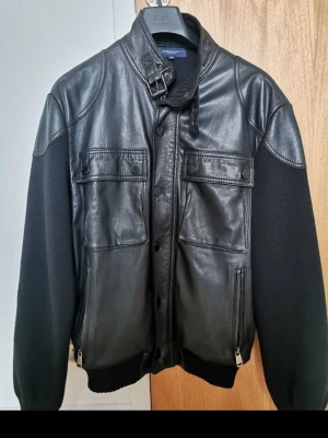 Burberry  - Snygg svart skinnjacka från Burberry med stickade ärmar och muddar. Jackan har två bröstfickor med lock och knappar, två sidofickor med dragkedja samt hög krage med spänne. Perfekt för dig som vill ha en unik och stilren look.