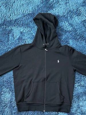 Ralph Lauren Zip up - Säljer en riktigt stilren Ralph Lauren Zip up i storlek M. Den är i färgen svart med en snygg grå logga, vilket ger ett väldigt rent och snyggt intryck. Tröjan har en grym passform och sitter precis som den ska.  Den är aldrig använd, så den är i nyskick och har inga tecken på slitage överhuvudtaget. Säljes då den tyvärr bara blivit liggande i garderoben.  Priset är inte hugget i sten och kan diskuteras vid en smidig affär.