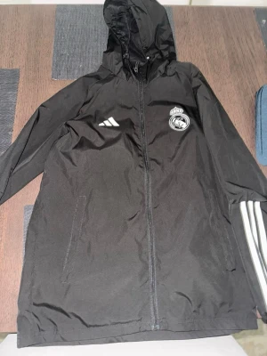 Svart Real Madrid vindjacka Adidas - Svart vindjacka från Adidas med Real Madrid-logga på bröstet och vita Adidas-ränder på ena ärmen. Jackan har huva, dragkedja framtill och två sidofickor. Perfekt för dig som vill ha sportig stil och visa ditt lagstöd.