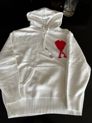 Ami Paris  - Säljer en vit hoodie från Ami paris med det ikoniska röda hjärta-A trycket på bröstet. Hoodien har huva med dragsko, magsko och ribbade muddar. Materialet är mjukt och stickat. Perfekt för en chill och stilren look. Är öppen för byten också. Hör av dig vid minsta fundering.