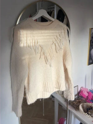 Krämvit stickad tröja med fransar - Säljer en krämvit stickad tröja från & Other Stories med fransdetaljer framtill. Tröjan har rund hals, långa ärmar och ribbade muddar. Perfekt för dig som gillar en mysig och trendig look.