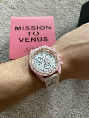 Omega x Swatch Mission to Venus klocka - Säljer en Omega x Swatch Speedmaster Mission to Venus från Bioceramic MoonSwatch-kollektionen. Klockan har en ljusrosa boett, vit urtavla med kronografer och vit rem med Omega-logga. Snygg och unik design som sticker ut på handleden.