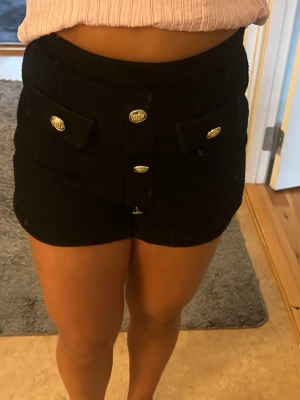 Svarta högmidjade shorts med guldknappar - Snygga svarta högmidjade shorts med dekorativa guldknappar framtill och två fickor med lock. Shortsen har en lite tjockare struktur och sitter tight. Perfekta för dig som vill ha en trendig och chic look. Köpt på Sellpy 