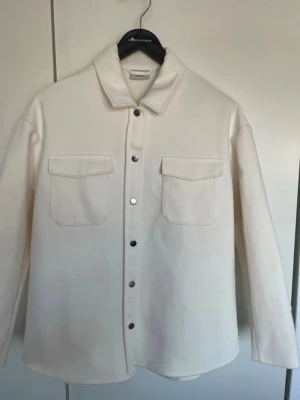 Vit overshirt med tryckknappar - Stilren vit overshirt med raka linjer, två stora bröstfickor med lock och silverfärgade tryckknappar framtill. Overshirten har klassisk krage och är tillverkad i ett mjukt, tjockare material som ger en clean look. Perfekt att bära öppen eller stängd.