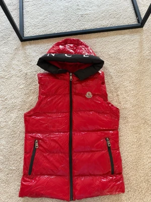 Röd dunväst från Moncler med huva - Snygg röd dunväst från Moncler med svarta detaljer. Västen har två dragkedjefickor framtill och Moncler-logga på bröstet. Perfekt för lager-på-lager och ger en sportig vibe. Lite slitage se på sista bilden