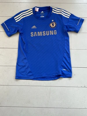 Chelsea FC matchtröja Adidas blå - Säljer en blå Chelsea FC matchtröja från Adidas med vita detaljer och tre ränder på axlarna. Tröjan har klubbmärke, Adidas-logga och Samsung som sponsortryck på bröstet. Tillverkad i lätt och ventilerande material med ClimaCool-teknologi.