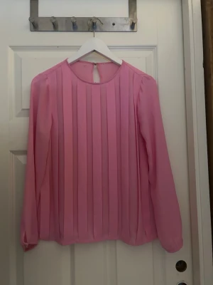 Rosa plisserad blus med lång ärm - Snygg rosa blus med plisserade detaljer framtill och rund halsringning. Blusen har långa ärmar med knapp vid ärmslut och en liten knapp i nacken. 