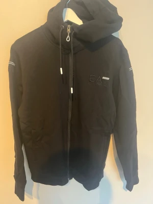 Svart hoodie från EA7 Emporio Armani - Svart hoodie med dragkedja från EA7 Emporio Armani. Den har huva med snören, diskret logotyp på bröstet och vita detaljer på ärmarna. Materialet är mjukt och bekvämt, perfekt för en avslappnad look. Långa ärmar och fickor på sidorna.