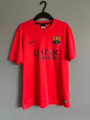Röd FC Barcelona Nike t-shirt - Säljer en röd FC Barcelona t-shirt från Nike med Qatar Airways-tryck på bröstet. Tröjan har klubbens emblem på vänster sida och Nike-logga på höger. Materialet är Dri-FIT, perfekt för sport och träning. Rund hals och korta ärmar.