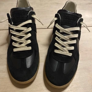 Svarta sneakers från Maison Margiela - Maison Margiela sneakers i svart läder och mocka med vita snören och gummisula. Klassisk design med diskret logotyp på tungan och vita sömdetaljer på hälen. Perfekt för dig som gillar stilrena och trendiga skor.
