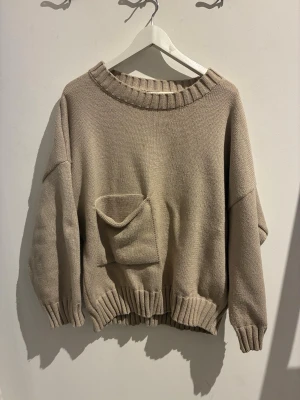 Beige oversized stickad tröja - Säljer en beige oversized stickad tröja med bred ribbad halsringning, muddar och nederkant. Tröjan har en stor ficka framtill och lös passform. Storleken är xxl men har funkat perfekt för mig som är S/M som en oversize tröja.
